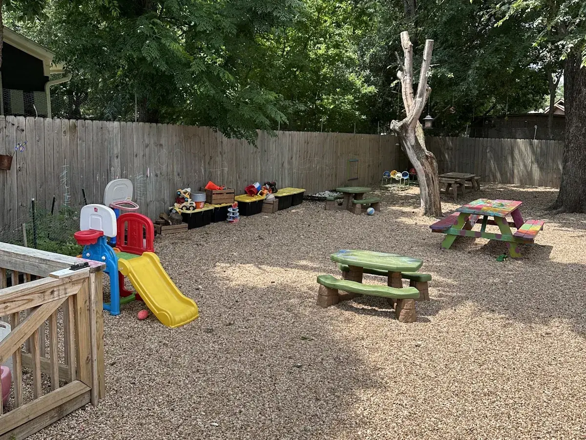 Fenced play yard under shade trees with picnic tables and a sandbox